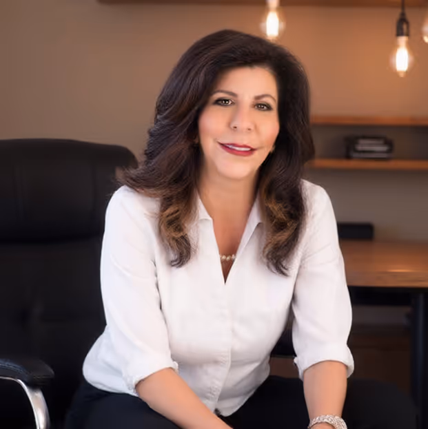 Woman with long dark hair wearing a white shirt sitting in a black office chair in a warmly lit office.