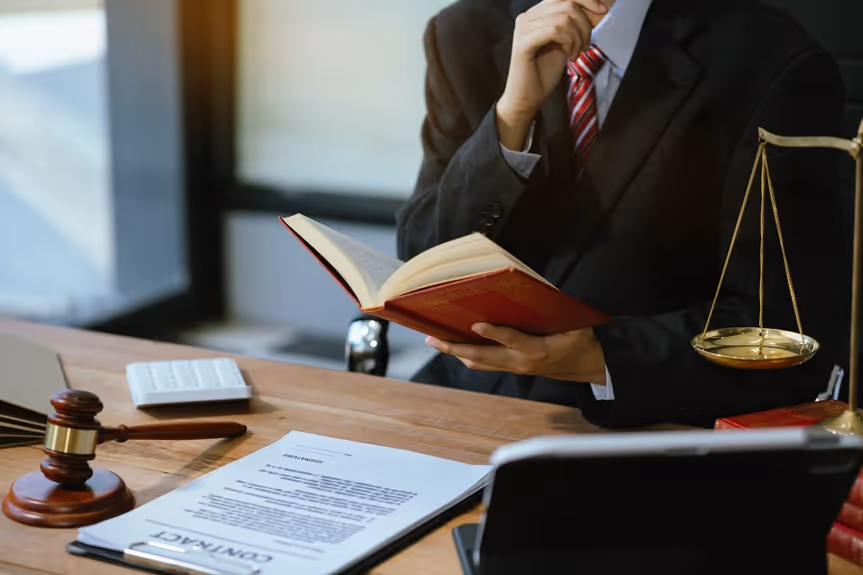 A person in a suit holding an open book at a desk with legal documents, a gavel, scales of justice, and a calculator.