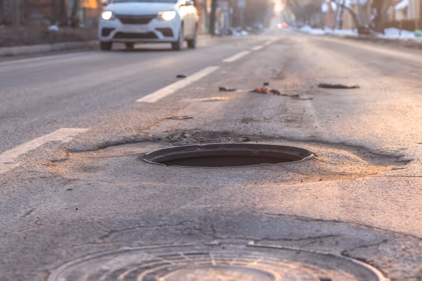 Open manhole cover creating a hazard in the middle of a street with a white car approaching in the background.