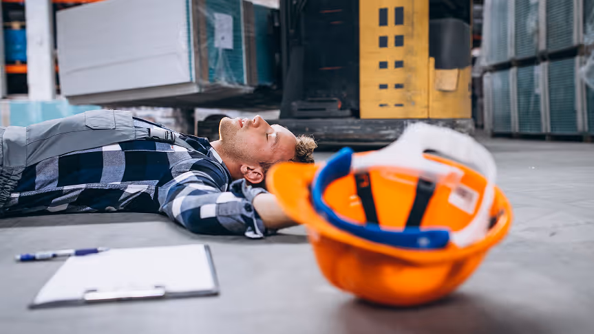 Construction worker lying unconscious on warehouse floor next to an orange safety helmet and clipboard.