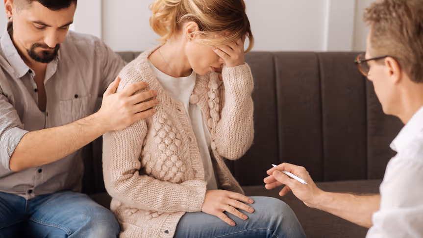 Woman sitting with her head in her hand looking distressed while a man comforts her and another person speaks to them.