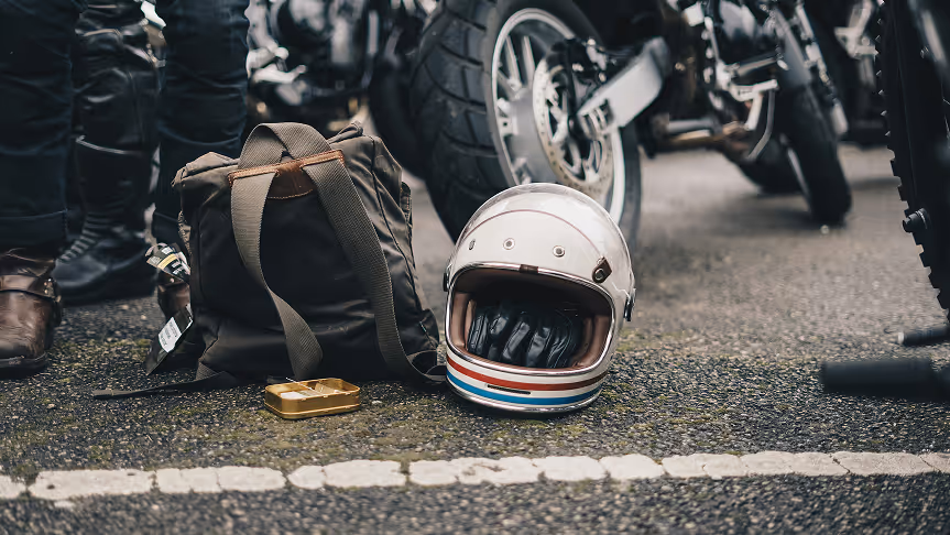 A vintage white motorcycle helmet with black gloves inside, a brown backpack, and a small gold tin on asphalt near parked motorcycles and people wearing black riding gear.