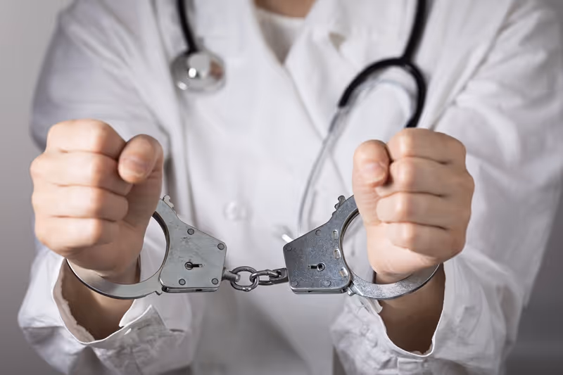 Doctor wearing a white coat and stethoscope with hands in metal handcuffs.