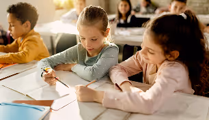 Two young girls sitting at a desk in a classroom, writing in notebooks, with other children in the background.