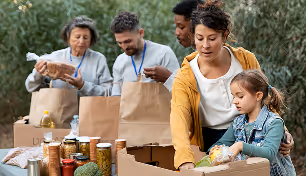 People packing food items into boxes outdoors, including a child and three adults organizing jars and bags.
