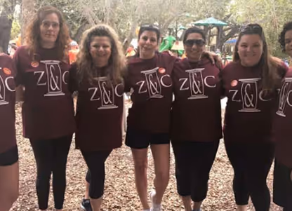 Group of six women standing outdoors with arms around each other, all wearing matching maroon shirts with 'ZINC' printed on them.
