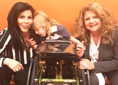 Two women smiling and posing with a young child who is resting on a walker, against an orange background.