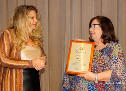 Two women facing each other; one holding a framed certificate or award, both smiling in a formal setting.