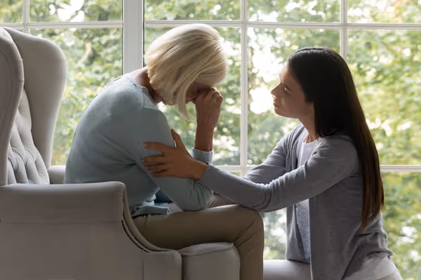 A young woman comforting an older woman who is sitting with her head in her hands by a window.