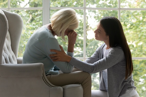 A young woman comforting an older woman who is sitting with her head in her hands by a window.
