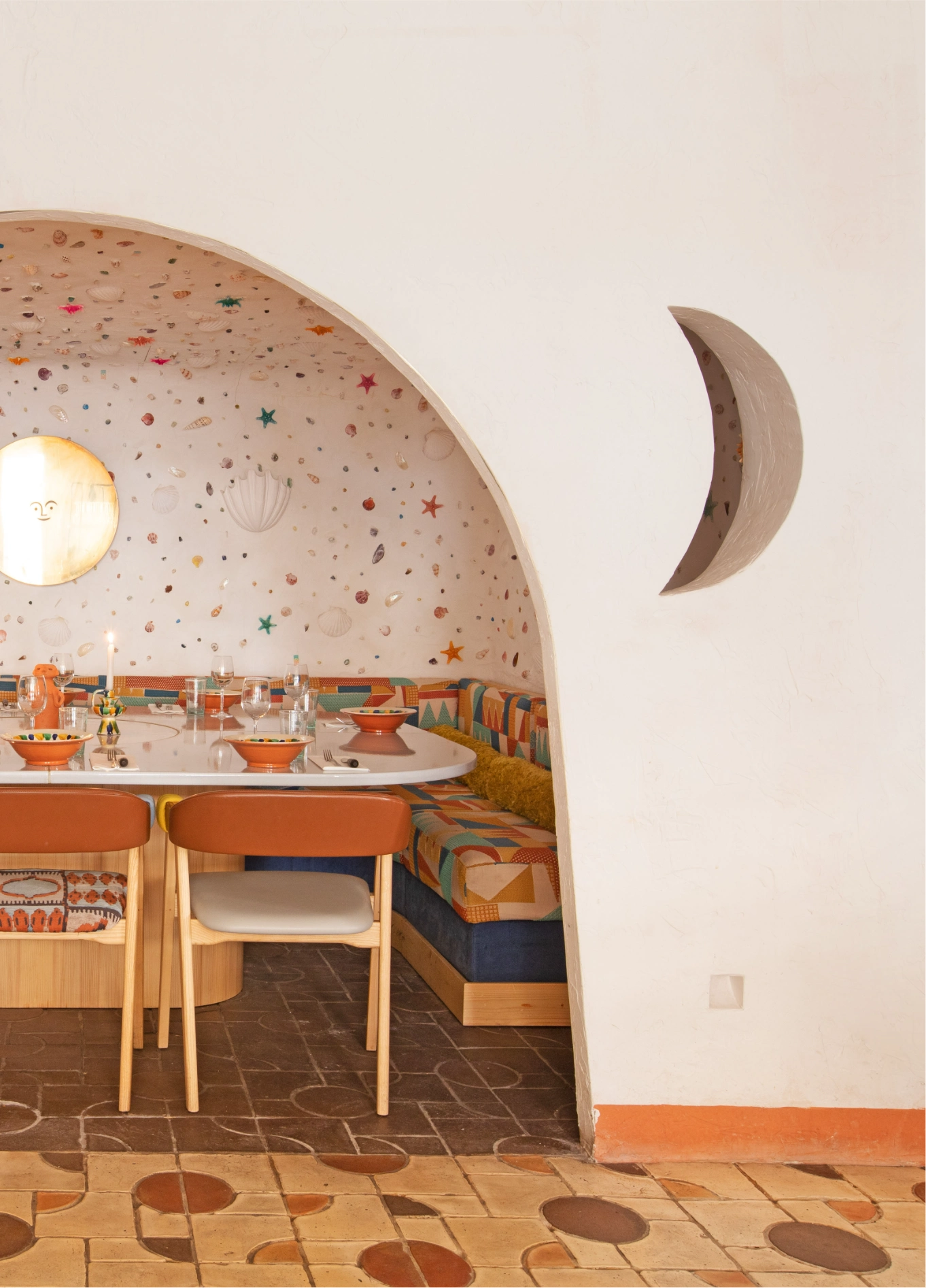 A dining area viewed through an archway, featuring a moon-shaped wall decoration, tables with chairs, and a tiled floor.