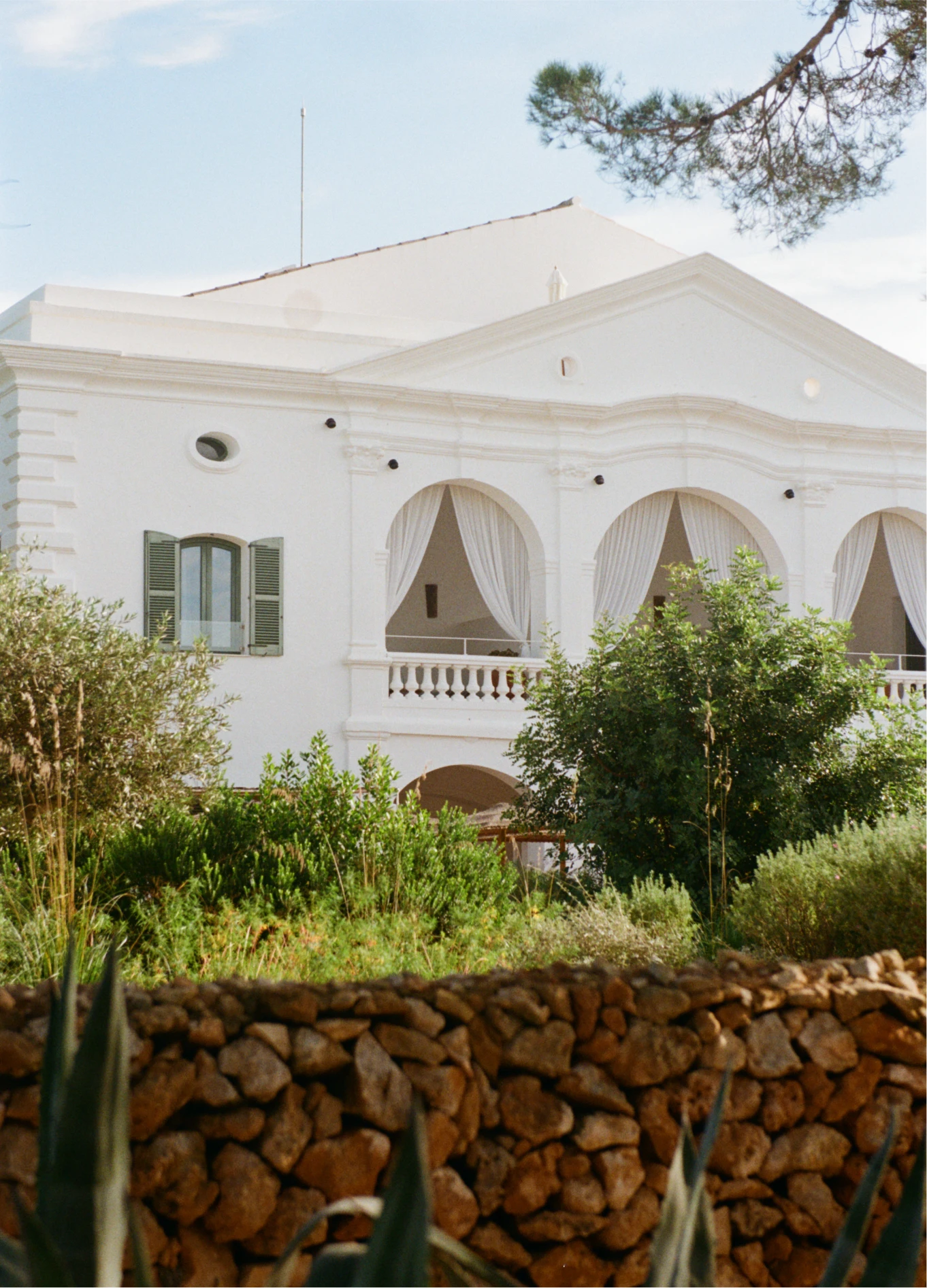 Immaculate white facade of the Menorca Experimental hotel, surrounded by lush vegetation and a dry stone wall in the foreground.