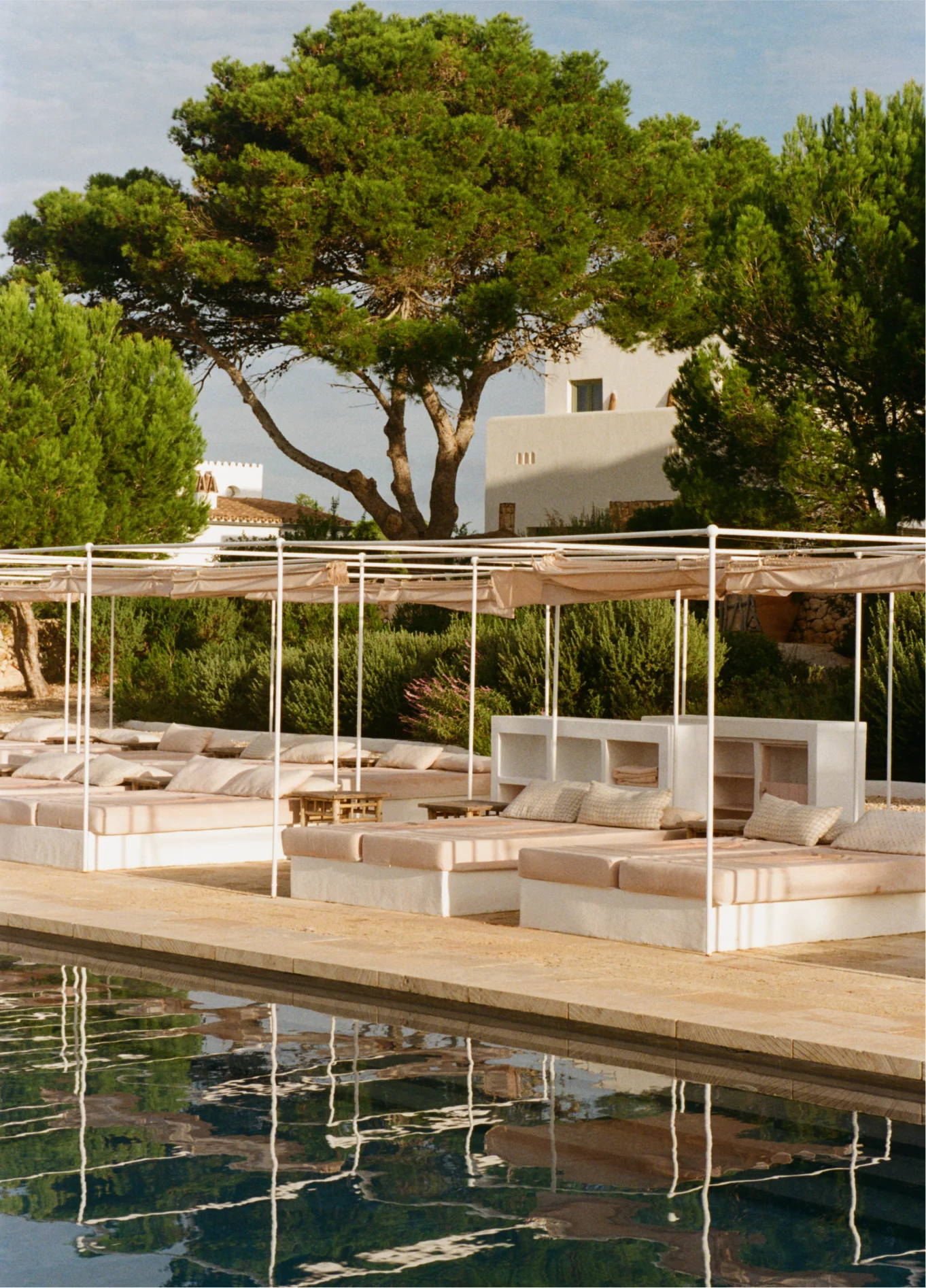 Outdoor swimming pool at Menorca Experimental with loungers under pergolas, surrounded by tall pine trees and a Mediterranean garden.