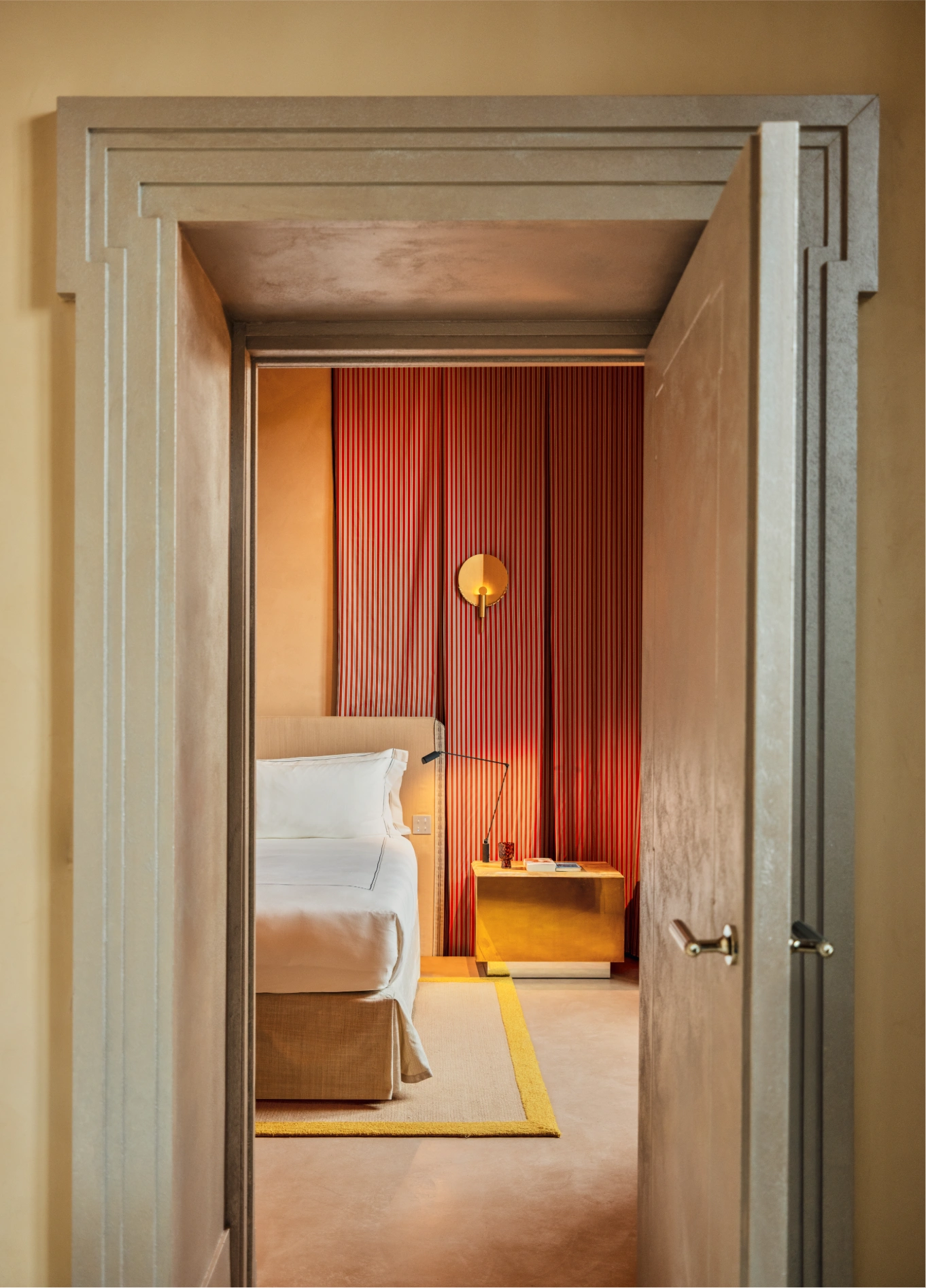 View through an ajar door leading into an elegant bedroom with warm tones, featuring a bed and a gold bedside table visible in the background.