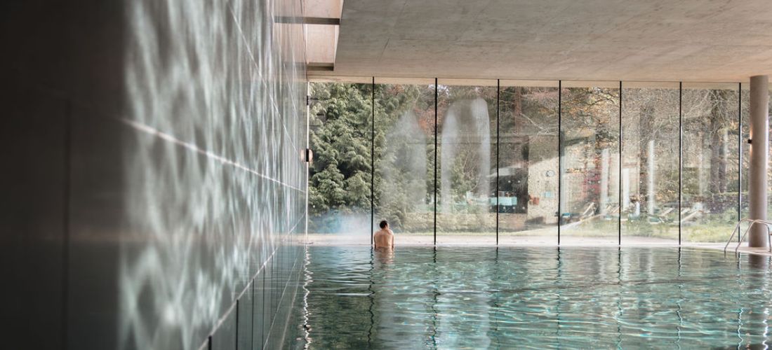 Serene indoor pool at Cowley Manor with glass walls overlooking greenery and fountains, reflecting calm light patterns.