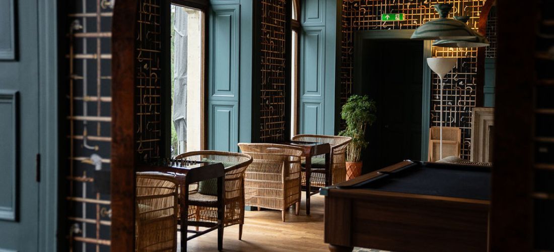 Elegante salón en Cowley Manor con sillas de mimbre, paneles de color verde azulado, paredes decorativas y una mesa de billar en un ambiente cálido y acogedor.