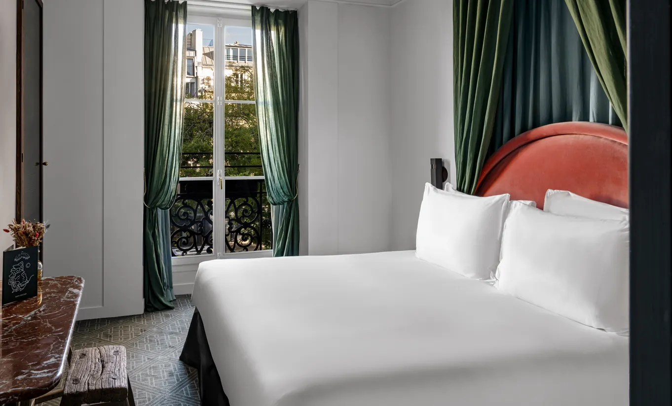 Chambre avec grand lit blanc, tête de lit rouge, rideaux vert sauge et fenêtre haussmannienne avec vue sur les toits parisiens dans la chambre Uzès de Grands Boulevards Experimental, hôtel boutique dans le 2e arrondissement de Paris