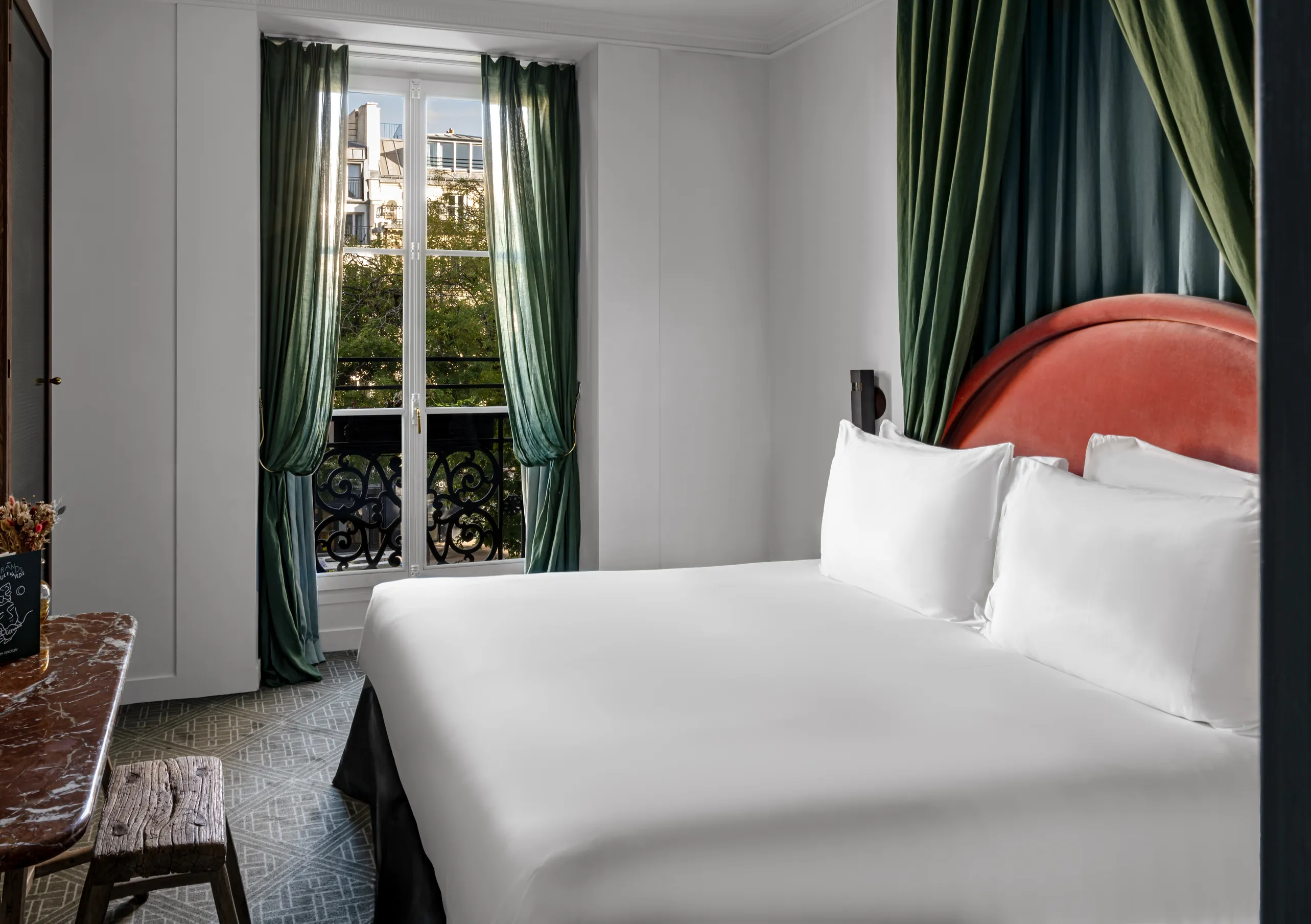 Chambre avec grand lit blanc, tête de lit rouge, rideaux vert sauge et fenêtre haussmannienne avec vue sur les toits parisiens dans la chambre Uzès de Grands Boulevards Experimental, hôtel boutique dans le 2e arrondissement de Paris