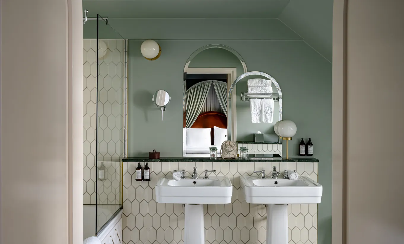 Salle de bain avec murs vert sauge, lavabo sur colonne et miroir rond sous plafond mansardé dans la chambre Sous les toits de Grands Boulevards Experimental, Paris 2e