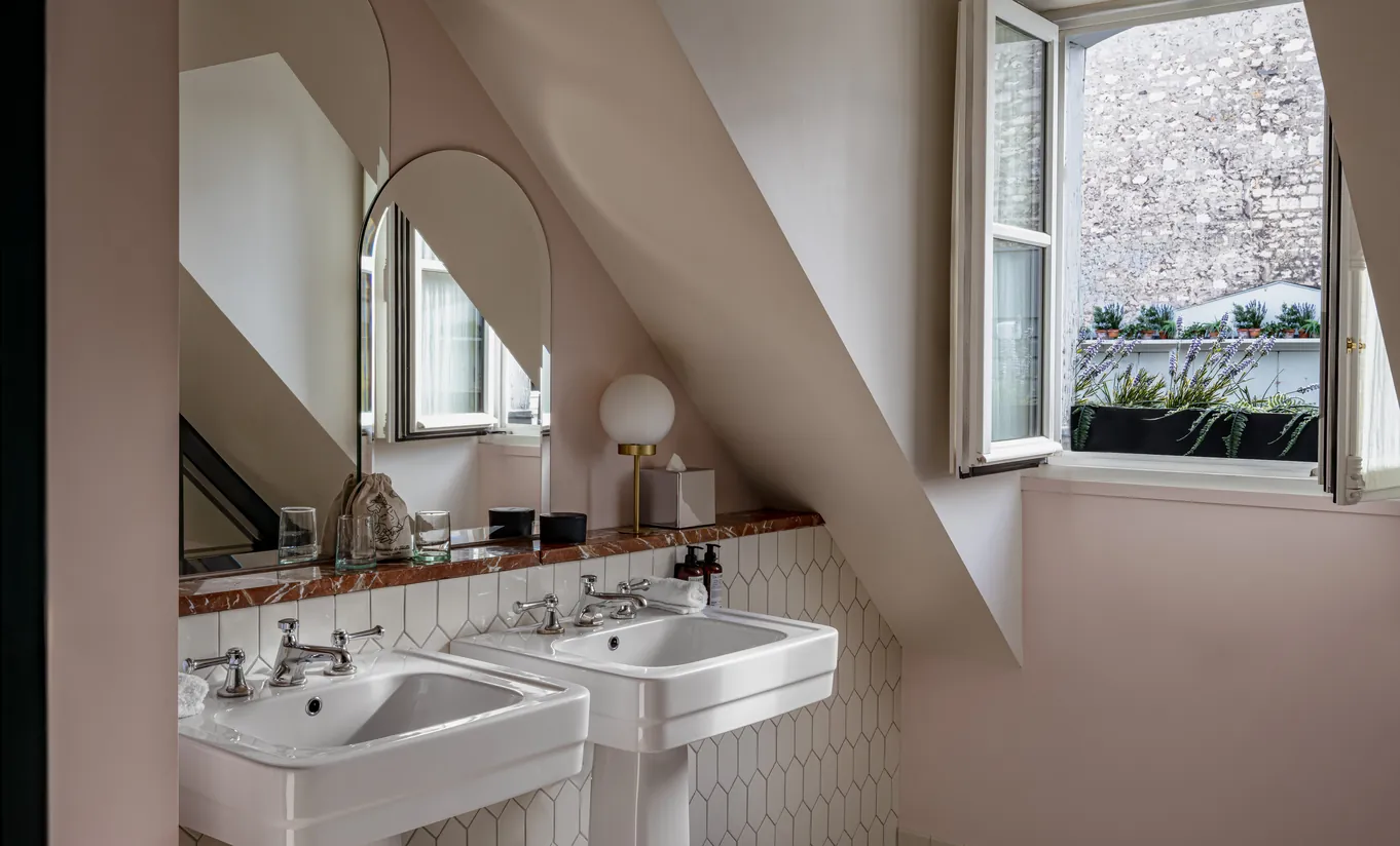 Salle de bain avec double lavabo sur colonne, poutres apparentes et fenêtre de toit sous les combles dans la chambre Sous les toits de Grands Boulevards Experimental, hôtel boutique de luxe sur les Grands Boulevards, Paris