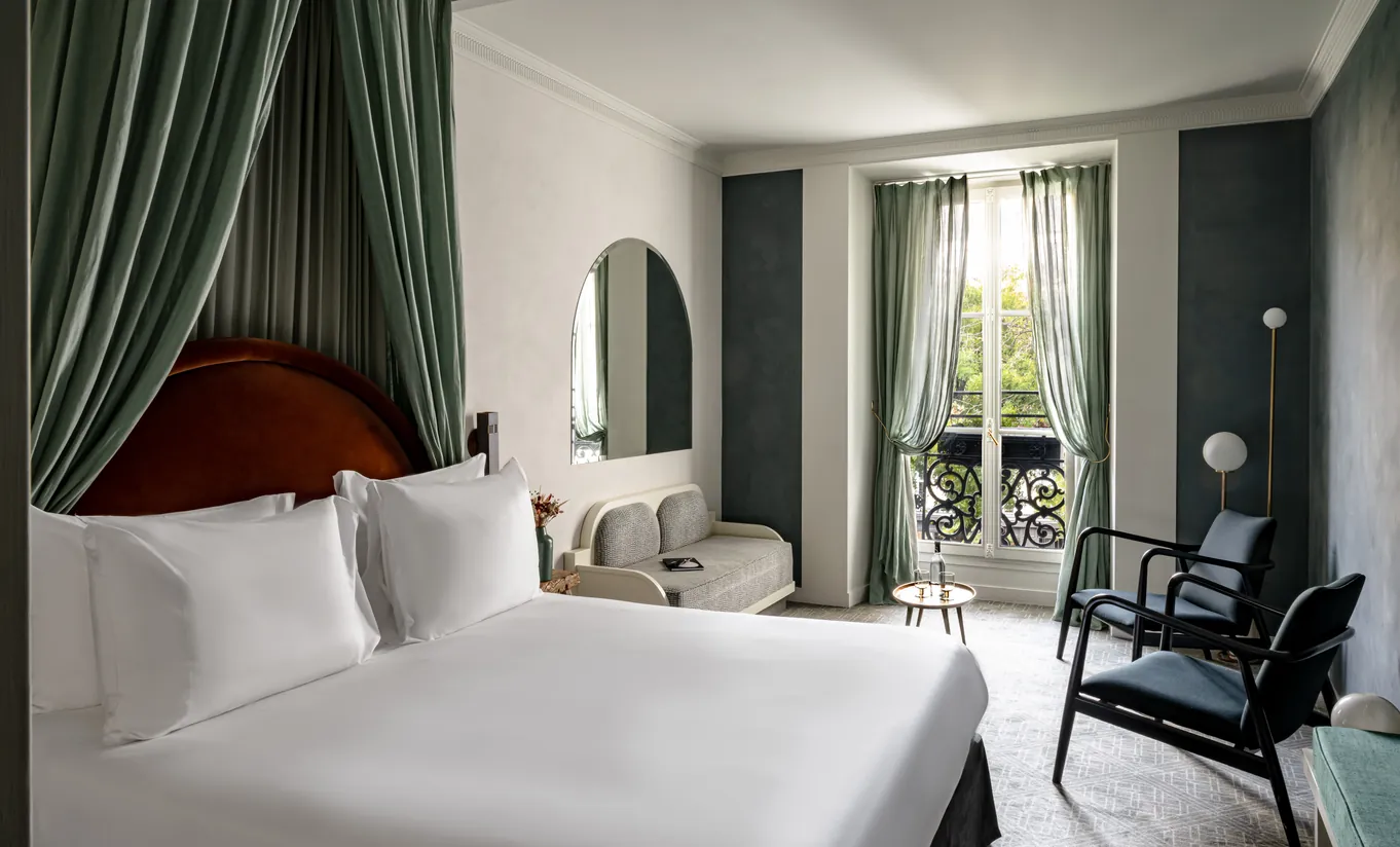 Chambre avec grand lit blanc, rideaux vert sauge, tête de lit rouge et vue sur les toits parisiens dans la chambre Grand Boulevard de Grands Boulevards Experimental, hôtel boutique dans le 2e arrondissement de Paris