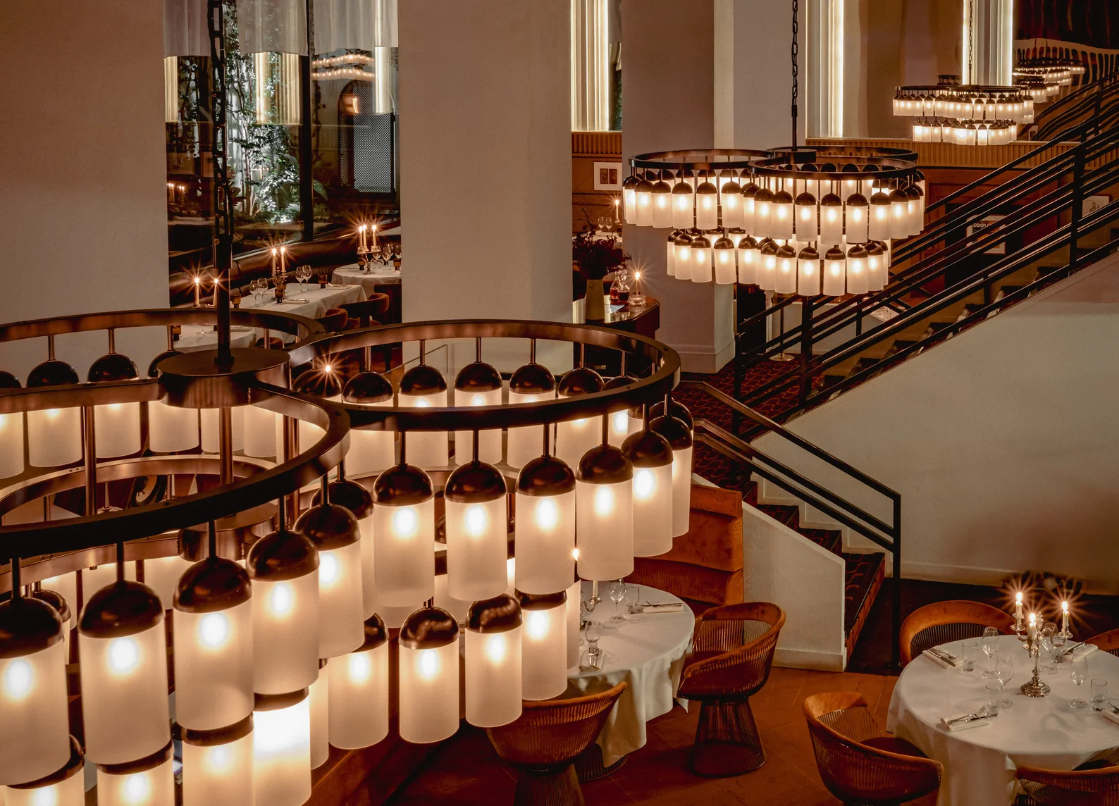 Comedor de doble altura con monumentales lámparas colgantes cilíndricas, escalera de caracol y mesas vestidas en blanco a la luz de las velas en el Hotel Experimental Marais de París