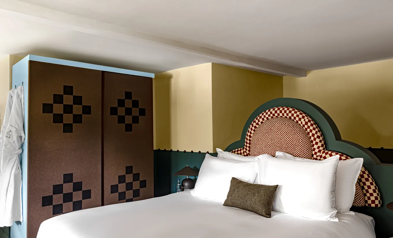 Chambre avec grand lit blanc, tête de lit en mosaïque, murs jaune moutarde et armoire à damier dans la chambre Wildflower de Cowley Manor Experimental, Cotswolds