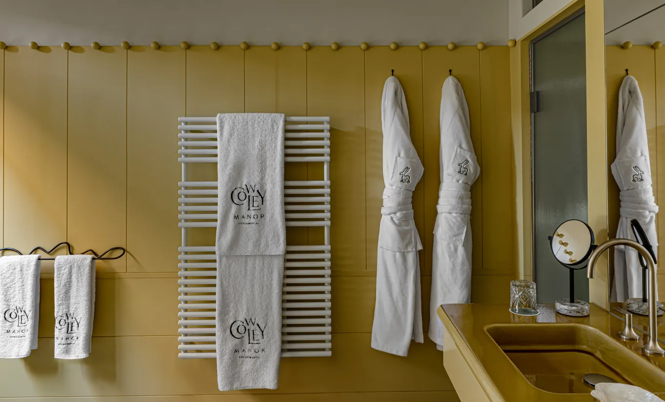 Deux peignoirs blancs sont suspendus à des crochets sur un mur jaune moutarde, à côté d'un sèche-serviettes blanc sur lequel est posée une serviette blanche brodée.