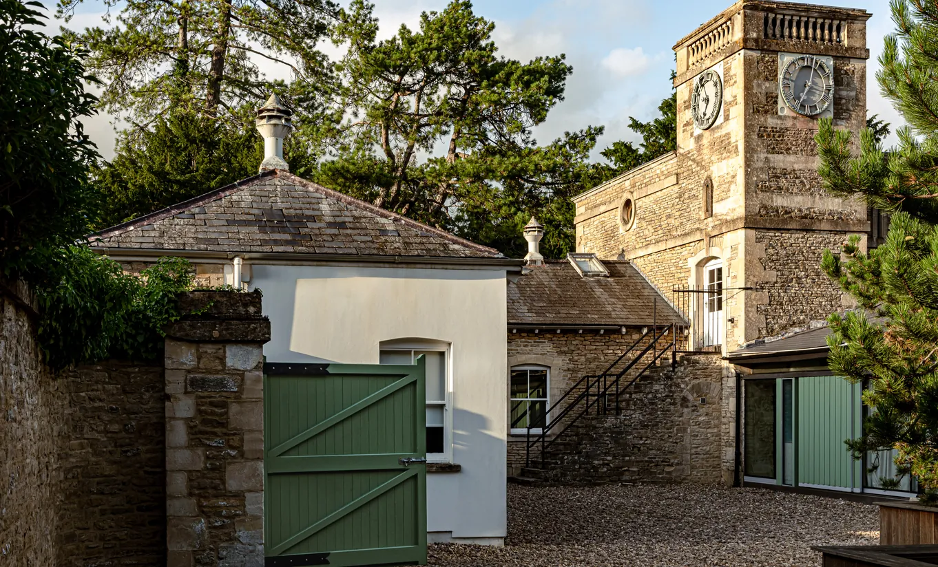 Vue extérieure de la tour de l'horloge en pierre, des dépendances du manoir et du portail vert à Cowley Manor Experimental, hôtel de luxe au cœur de la campagne des Cotswolds.