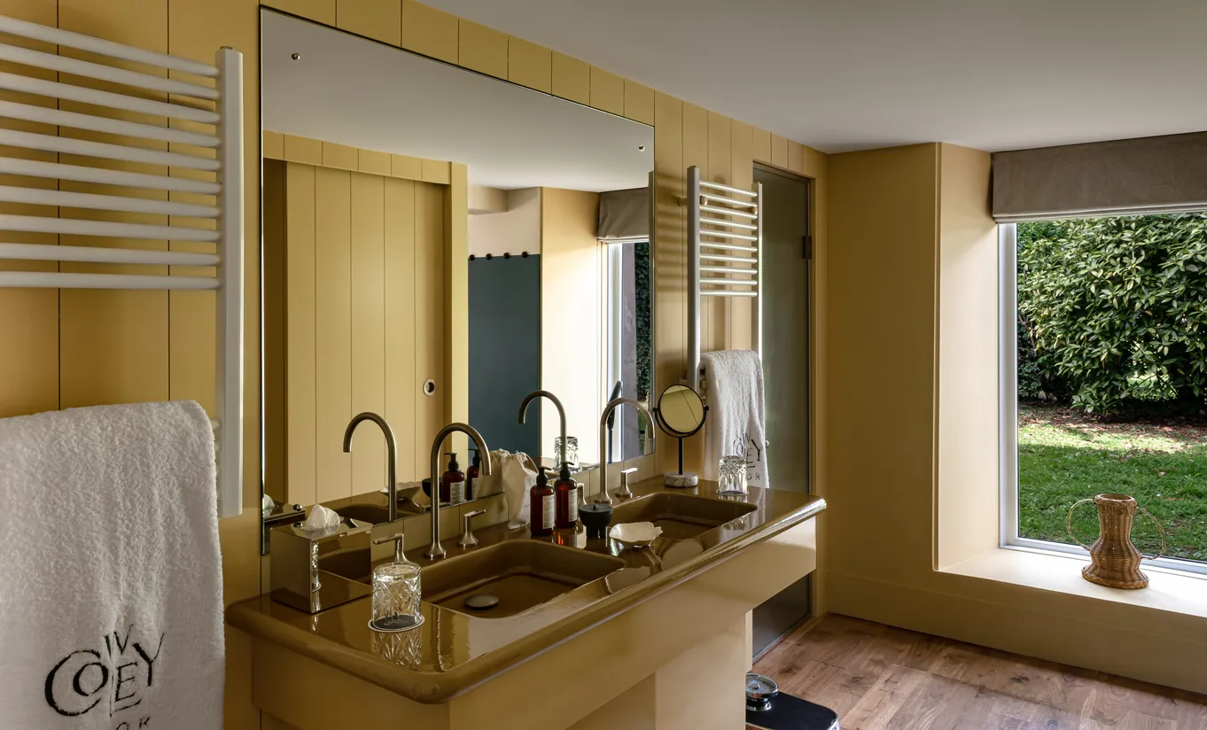 Salle de bain avec murs jaune moutarde, meuble vasque en bois et fenêtre donnant sur le jardin dans la chambre Wildflower avec terrasse de Cowley Manor Experimental, Cotswolds