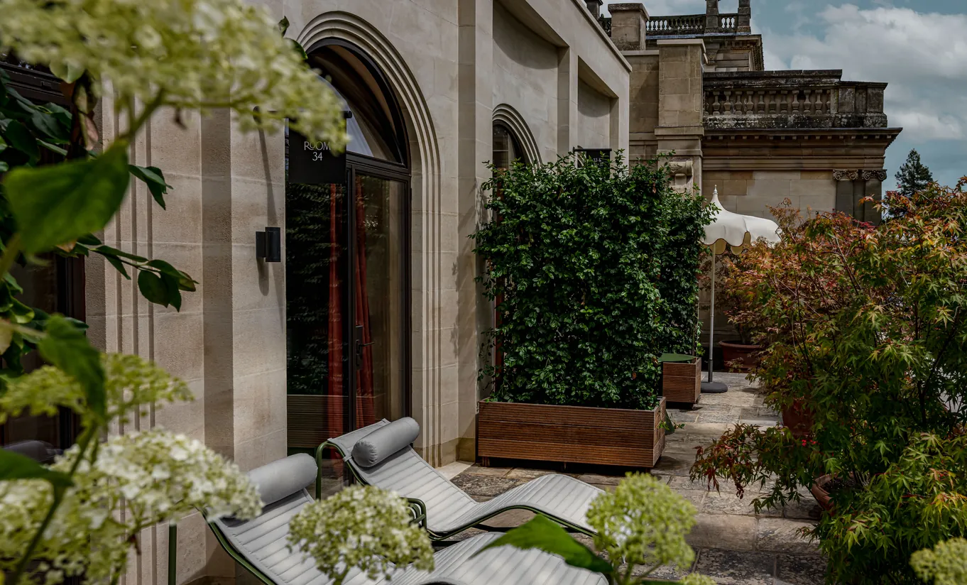 Vue extérieure de la façade en pierre du manoir avec une terrasse privée, un salon de jardin et des hortensias à Cowley Manor Experimental, dans les Cotswolds.