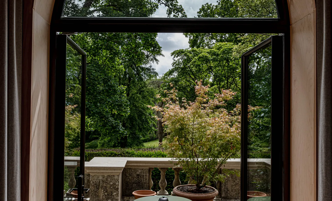 Fenêtre en arche ouvrant sur une terrasse privée verdoyante entourée d'arbres dans la chambre Waterlily with Terrace de Cowley Manor Experimental, idéal pour une escapade à la campagne dans les Cotswolds