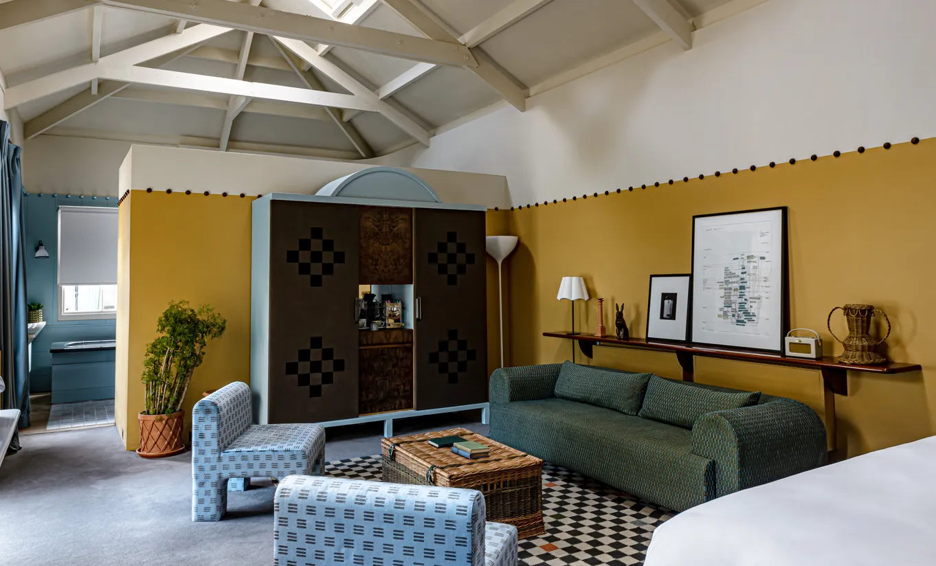 Chambre spacieuse avec plafond voûté, murs jaune moutarde, canapé vert et tapis à motifs dans la Churns River Junior Suite de Cowley Manor Experimental, retraite à la campagne dans les Cotswolds