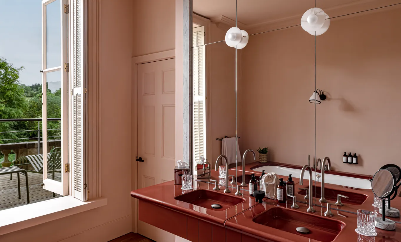 Salle de bain lumineuse avec murs terracotta, double vasque rouge et fenêtre ouverte sur le jardin dans la St Mary Suite de Cowley Manor Experimental, Cotswolds