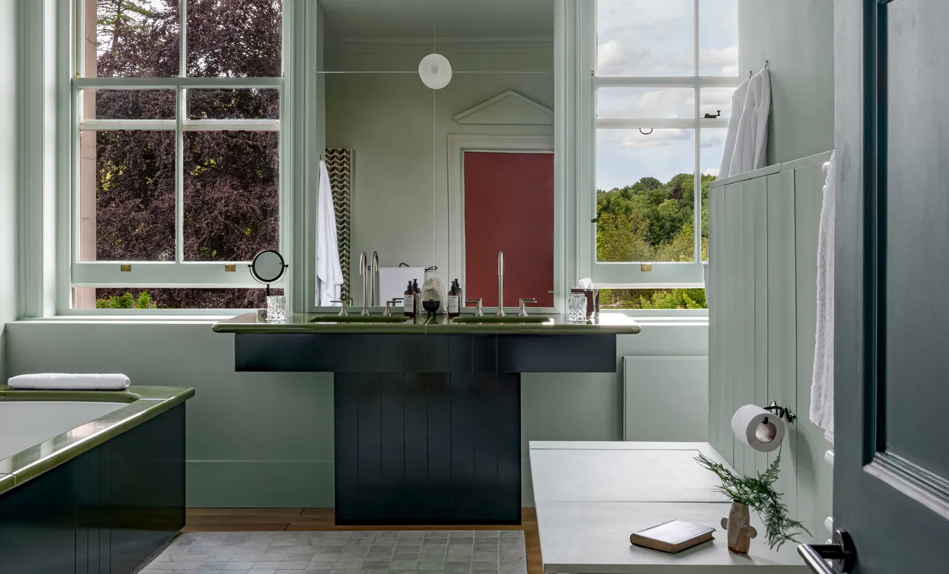 Salle de bain lumineuse avec meuble vasque vert sauge, double lavabo et fenêtre donnant sur la campagne des Cotswolds dans la Swan Lake Suite de Cowley Manor Experimental