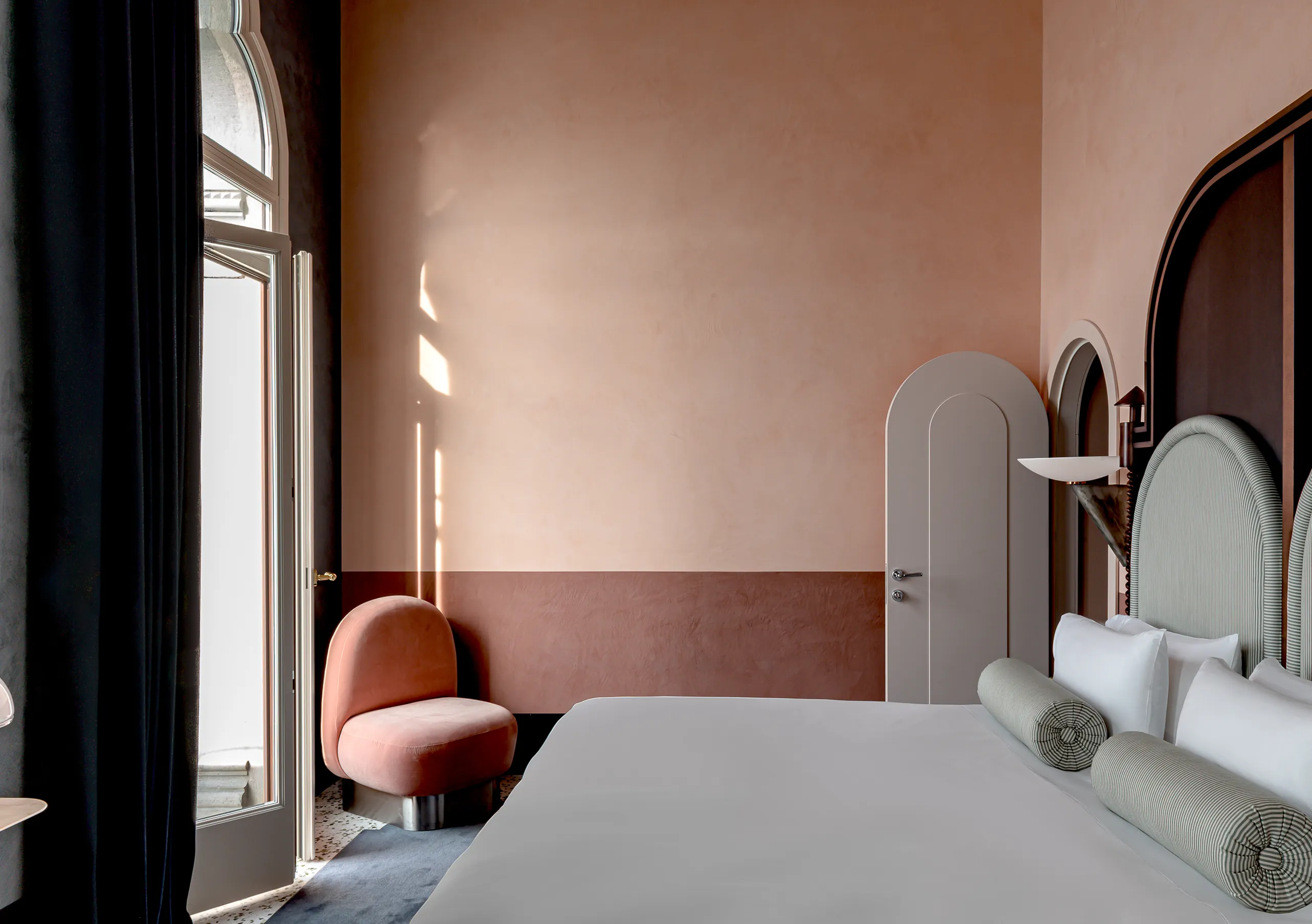 Il Palazzo Experimental bedroom with tall arched window, blush pink walls, velvet armchair and striped headboard in Venice.