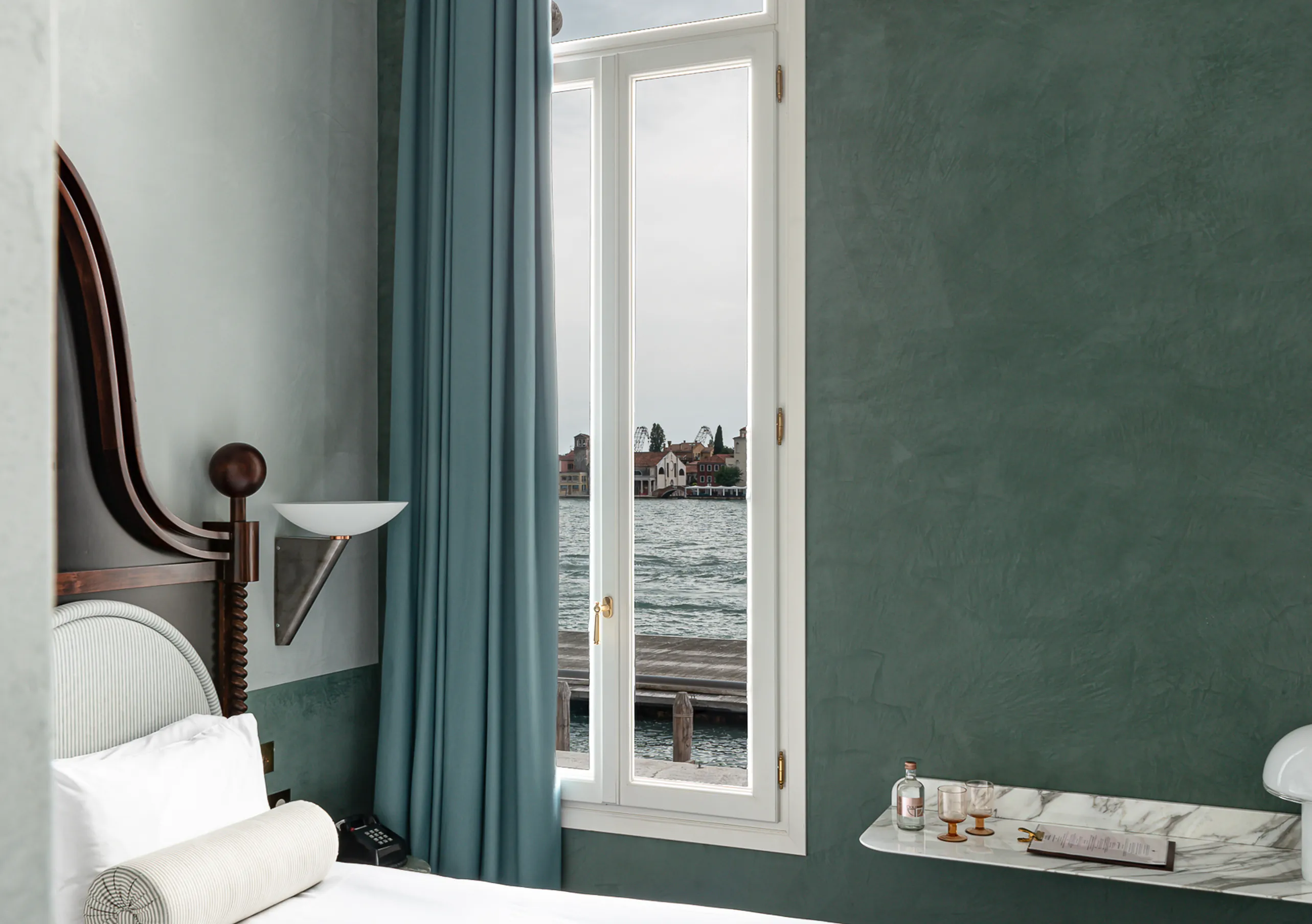 Il Palazzo Experimental hotel room with teal walls, open lagoon window view and marble drinks shelf in Venice.