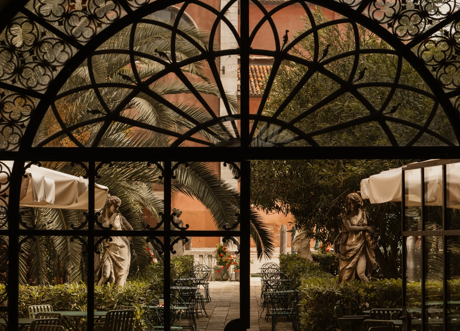 Vista attraverso una finestra in ferro battuto sul giardino veneziano con statue e palme al Giardino Ognissanti, Il Palazzo Experimental, Venezia.