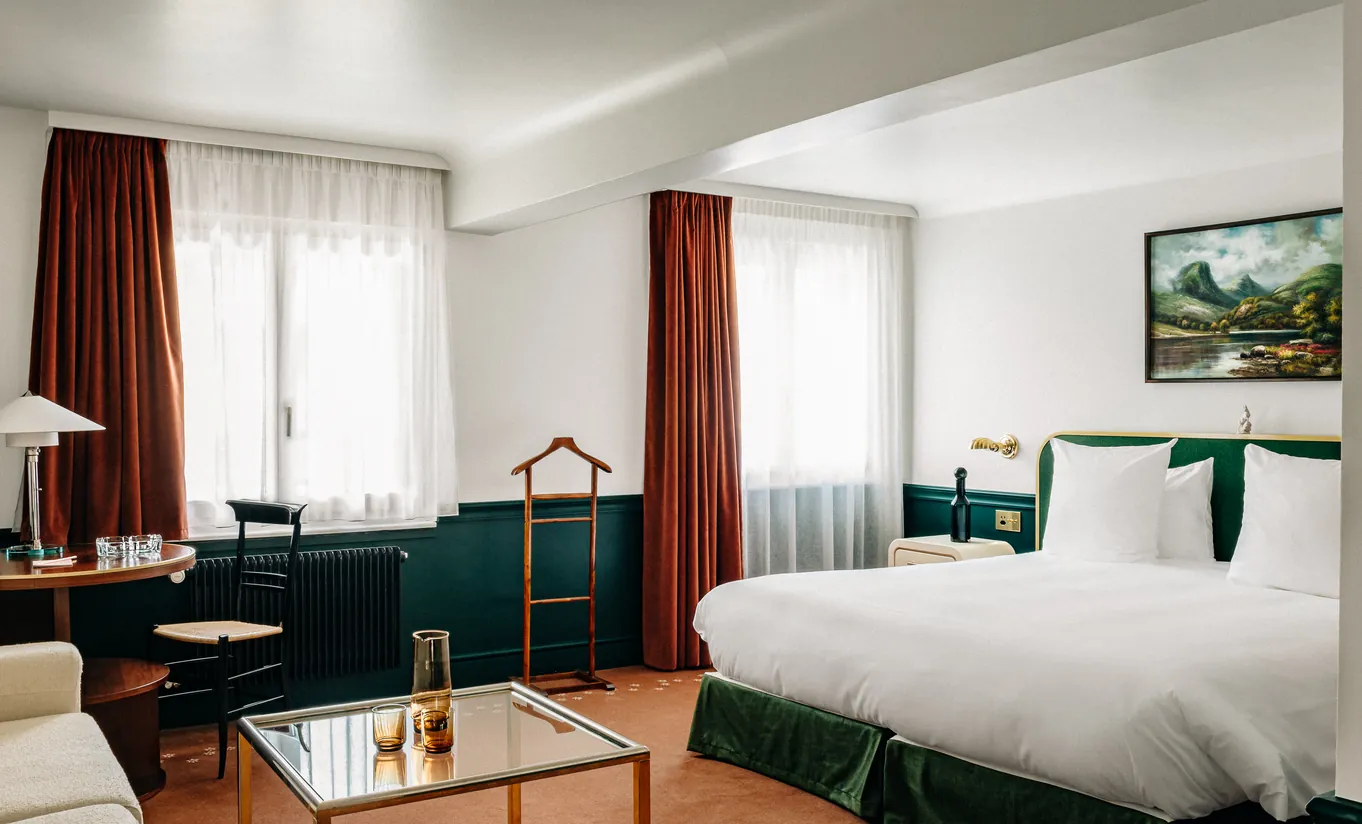 Bright bedroom with a white double bed, green armchair and red curtains in the Junior Suite at Experimental Chalet Verbier