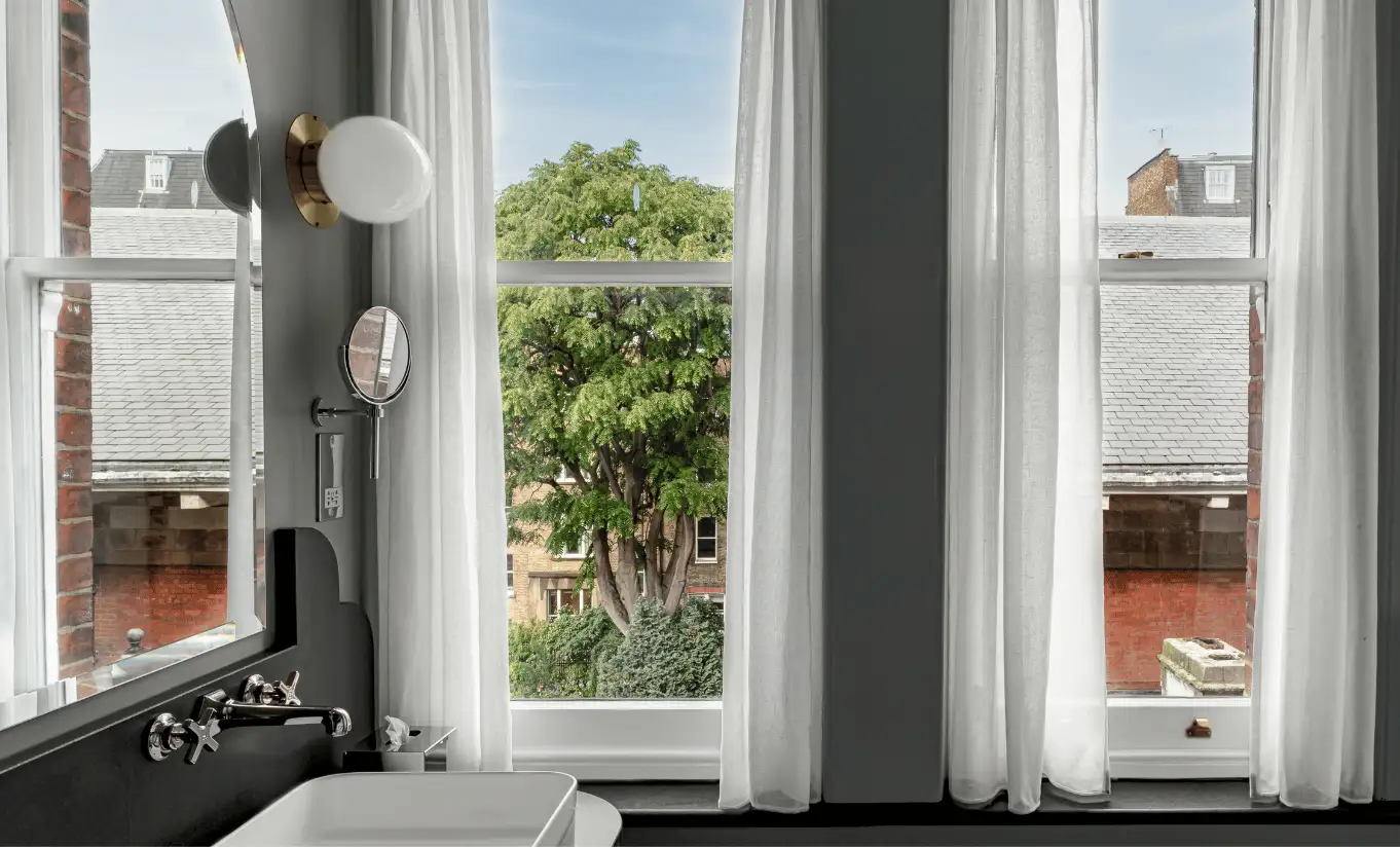 Bright bathroom with a freestanding bathtub, large sash windows and a view of the rooftops in the Saint Paul Executive Room at Henrietta Experimental, luxury boutique hotel in central London