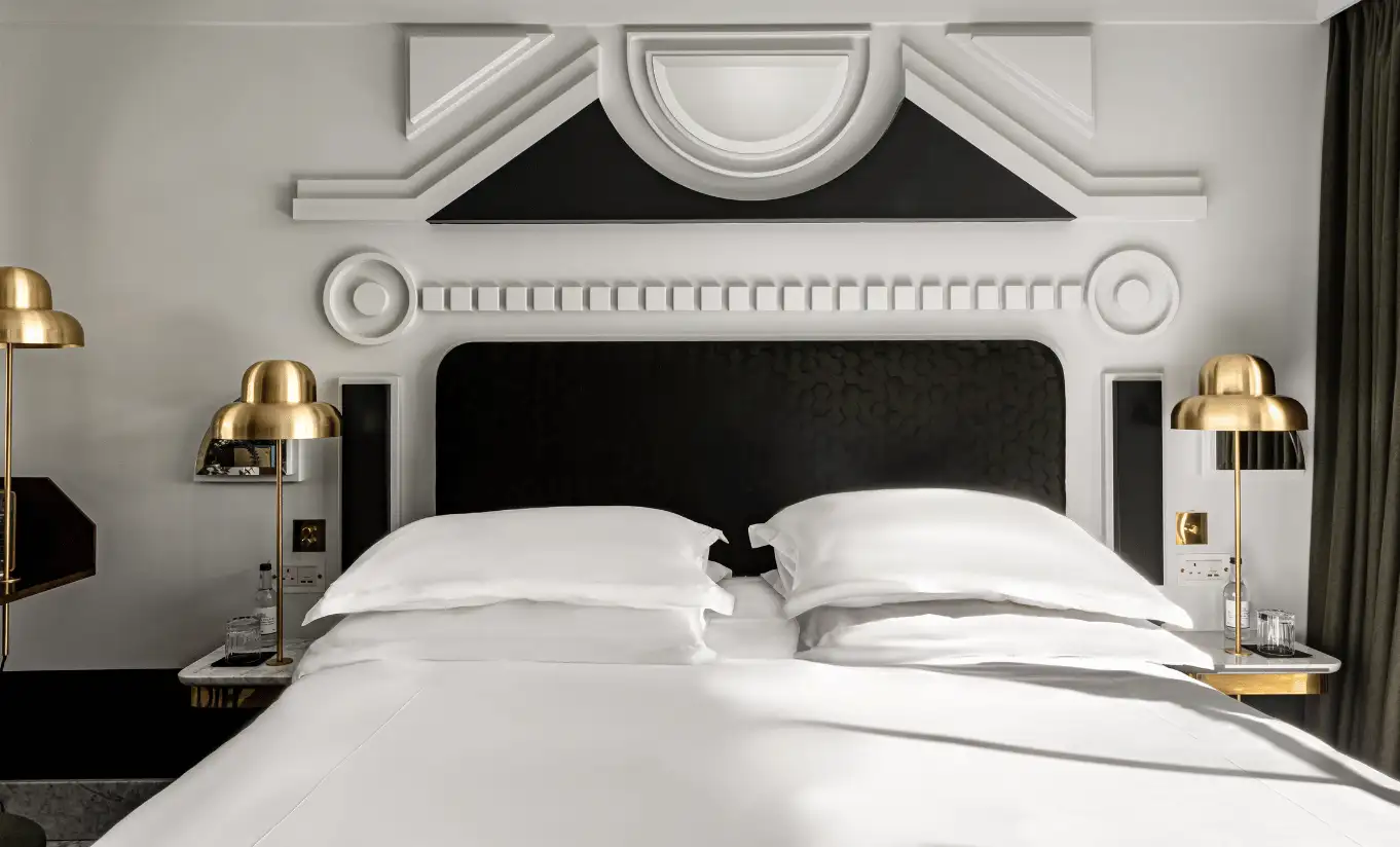 Detail of a white double bed with a black upholstered headboard and gold wall lights in the London Eye Superior Terrace room at Henrietta Experimental, luxury boutique hotel in central London