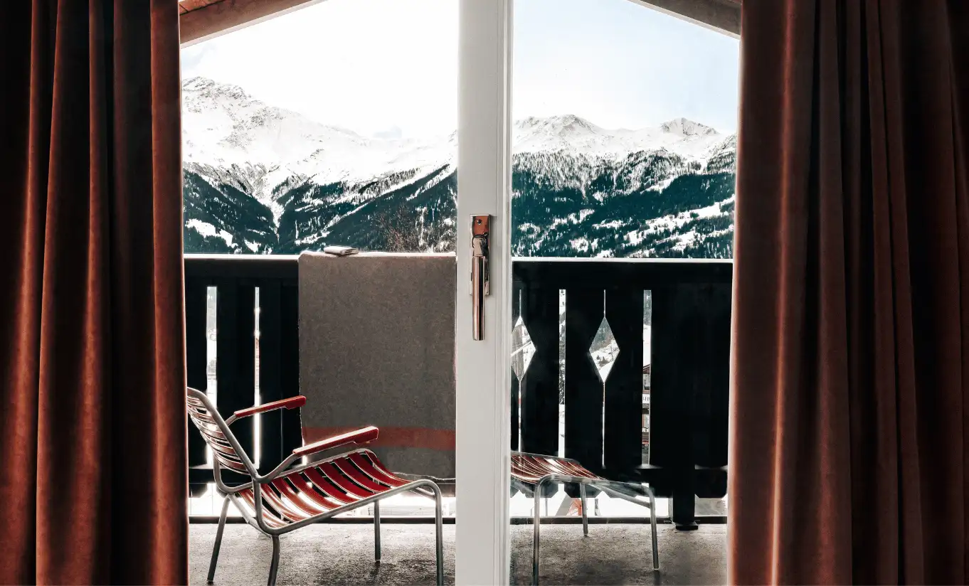 Balcony with red chairs and floor-to-ceiling windows overlooking the snowy Swiss Alps in the Panorama Suite at Experimental Chalet Verbier