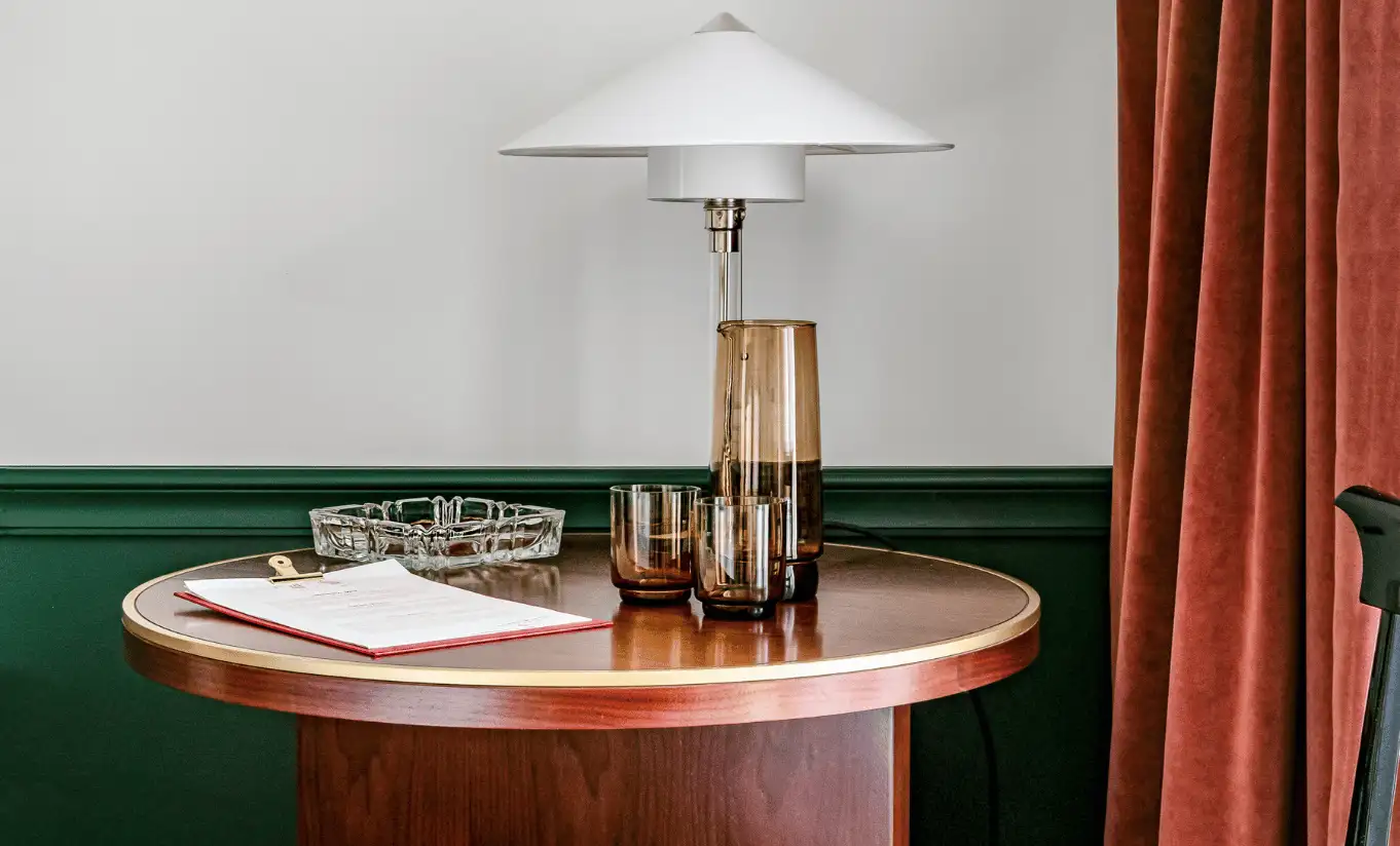 Detail of a bedroom with a round wooden side table, a lamp and dark green wall panelling in the Classic Room at Experimental Chalet Verbier