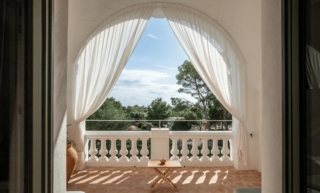 Private balcony with sheer white curtains billowing in the breeze and lush Mediterranean landscape views at Menorca Experimental hotel, Experimental Group.