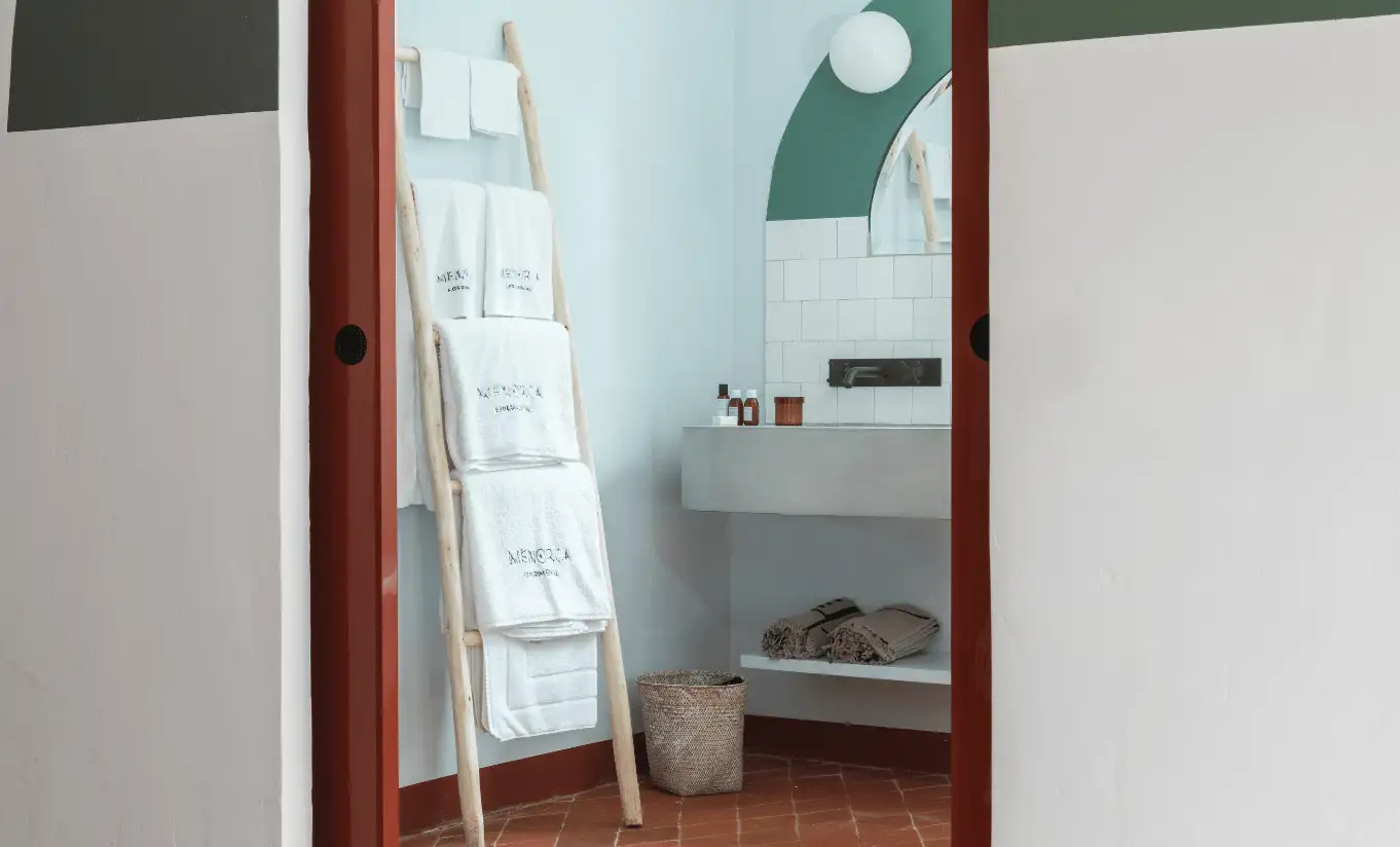 Minimalist bathroom glimpsed through a doorway with wooden ladder towel rack and stone basin at Menorca Experimental hotel, Experimental Group.
