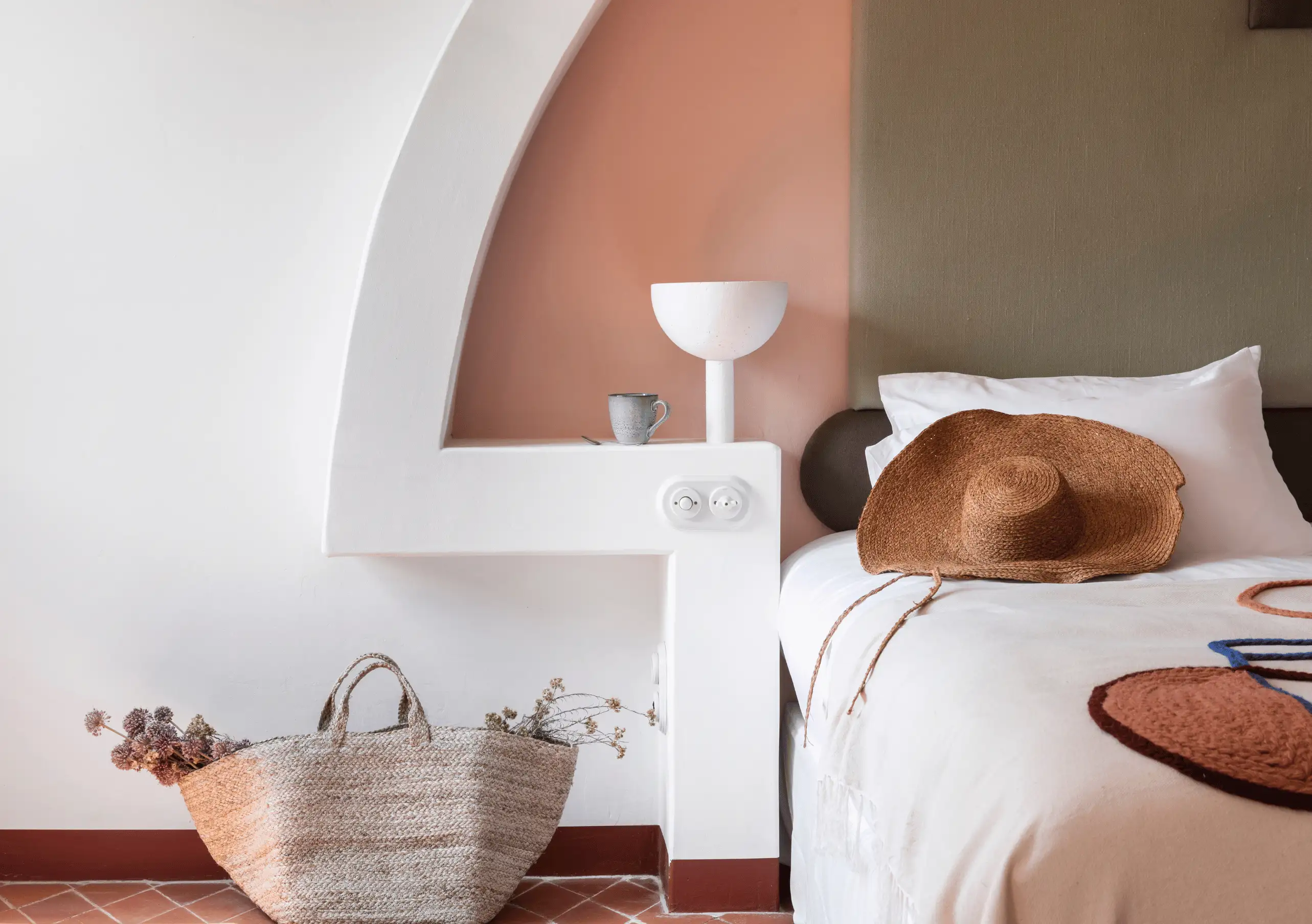 Relaxed guest lounging on white bedding beneath a terracotta arched niche with woven bag and bedside accessories at Menorca Experimental hotel, Experimental Group.