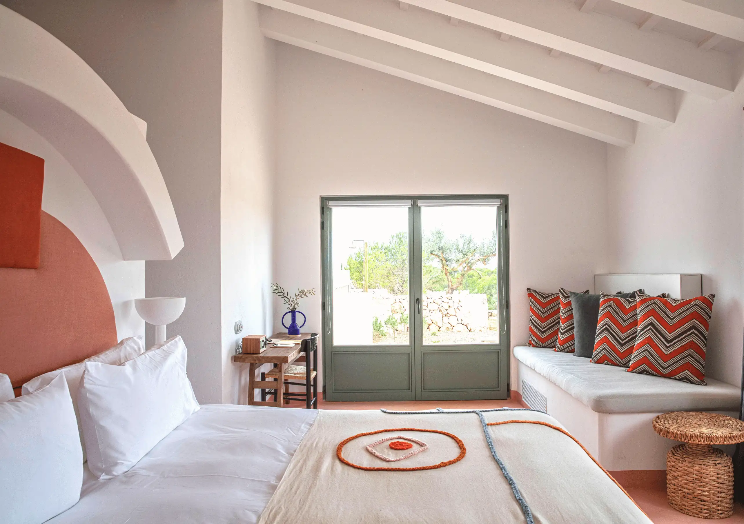 Menorca Experimental hotel bright bedroom with terracotta headboard, white bedding and garden view through large windows.