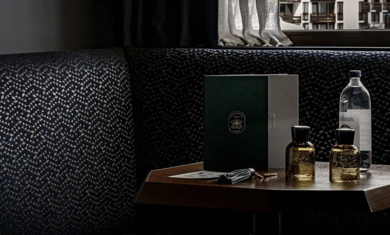 Moody bedside vignette with dark leather upholstery, a glass candle holder, perfume bottle and green hardcover book at Experimental Chalet Verbier, Experimental Group.