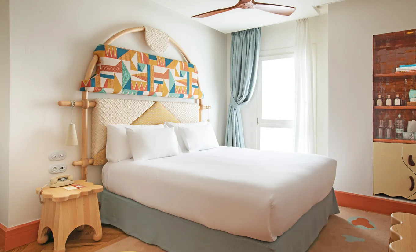 Whimsical bedroom with an ornate arched headboard, white linen bedding, wooden side table and teal curtains at Montesol Experimental hotel, Experimental Group.