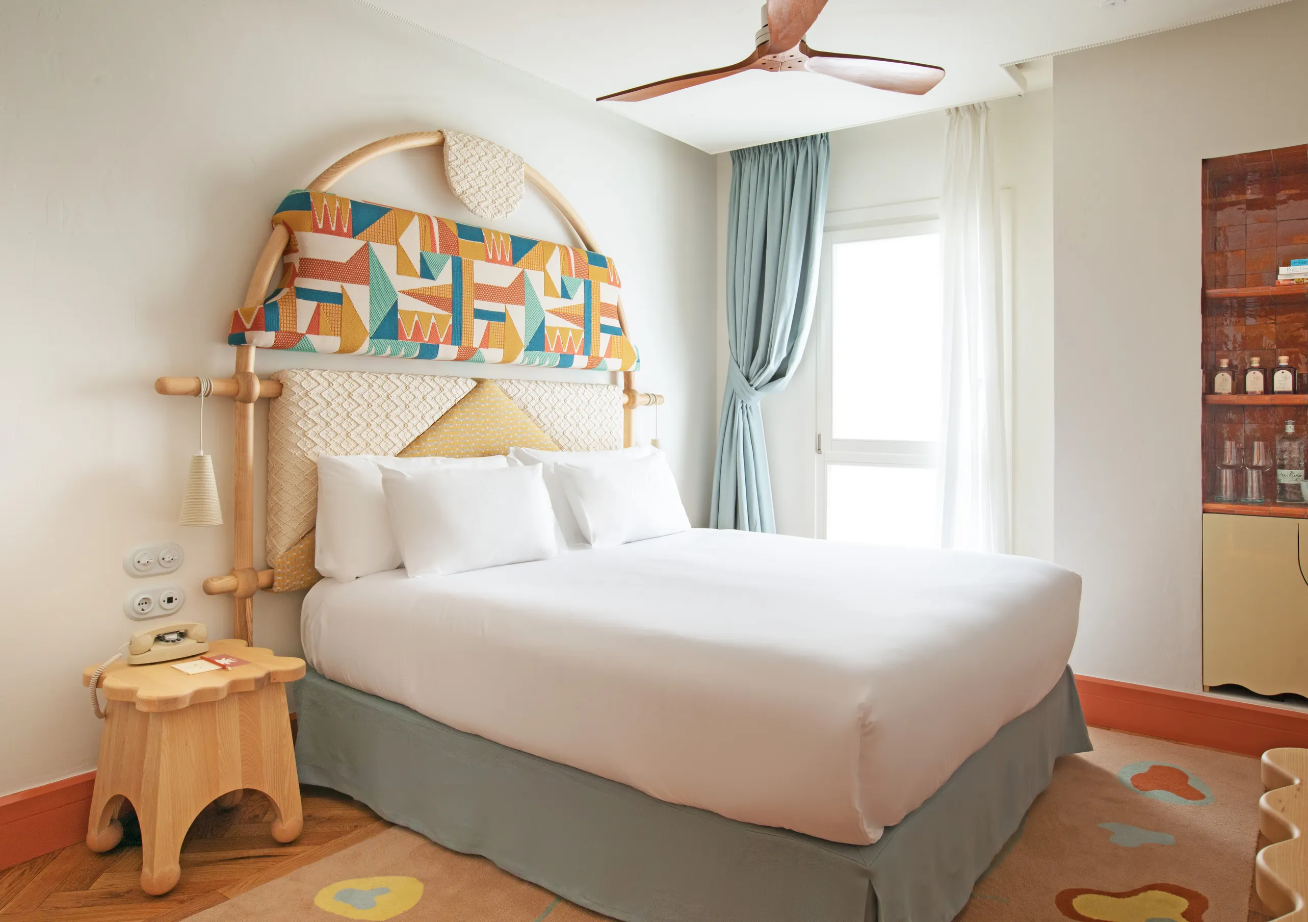 Charming hotel bedroom with a decorative arched headboard, soft white linen, teal window curtains and ceiling fan at Montesol Experimental hotel, Experimental Group.
